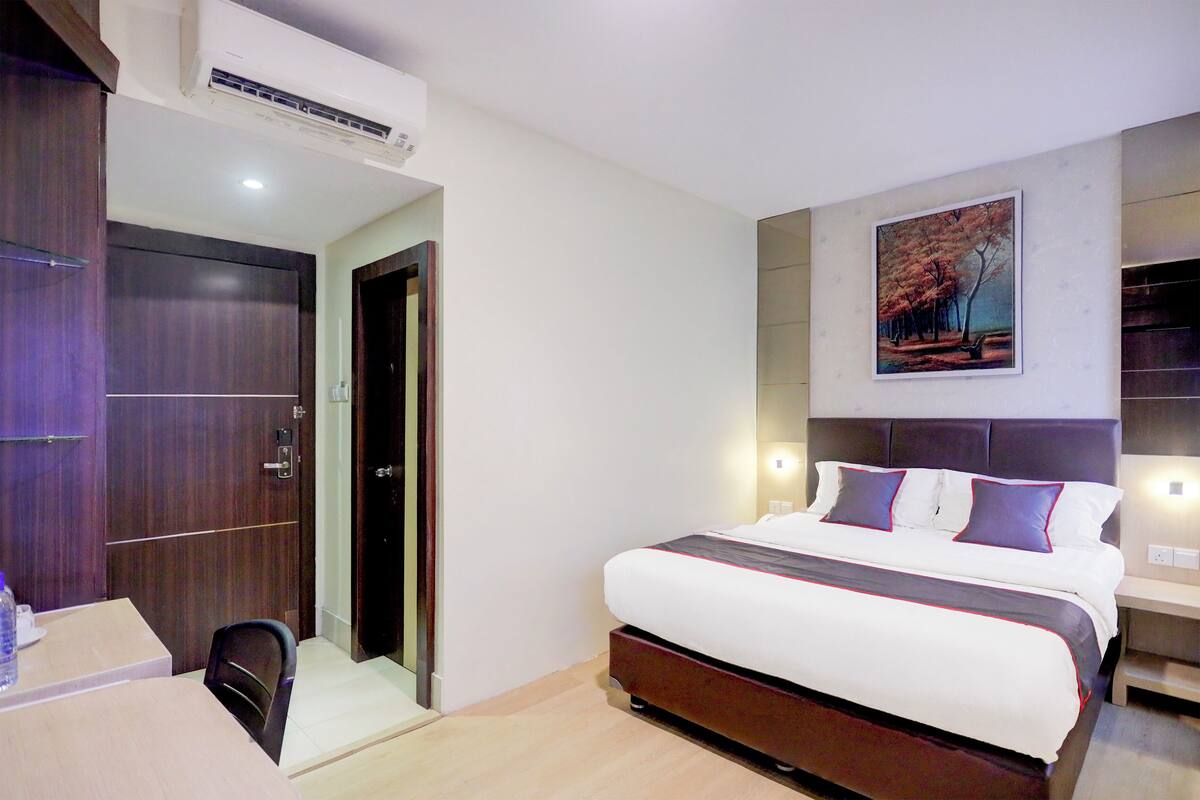 A hotel room with a large bed, a desk, and a chair. There is a painting on the wall and an air conditioning unit above the bed.