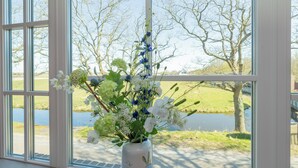 Een kamer met grote ramen, een vaas met bloemen, en uitzicht op een meer en bomen.