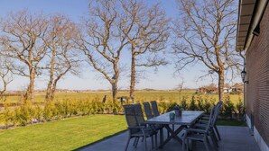 Een terras buiten met een tafel en stoelen, omringd door bomen en uitzicht op een veld.