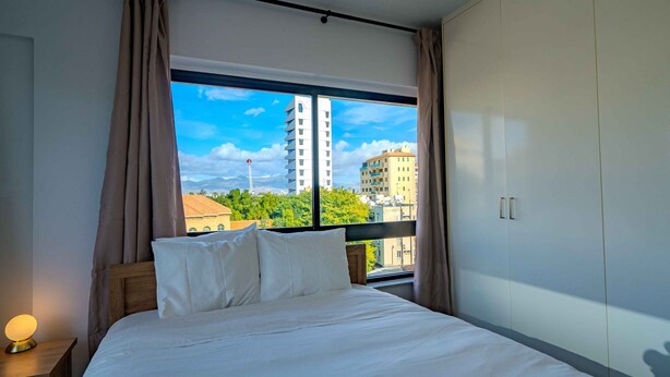 Una camera d'albergo con un letto grande, un comodino con una lampada e vista su edifici e alberi attraverso una finestra.