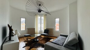 A modern living room with a glass coffee table, a grey sofa, a black chair, and a unique ceiling fan.