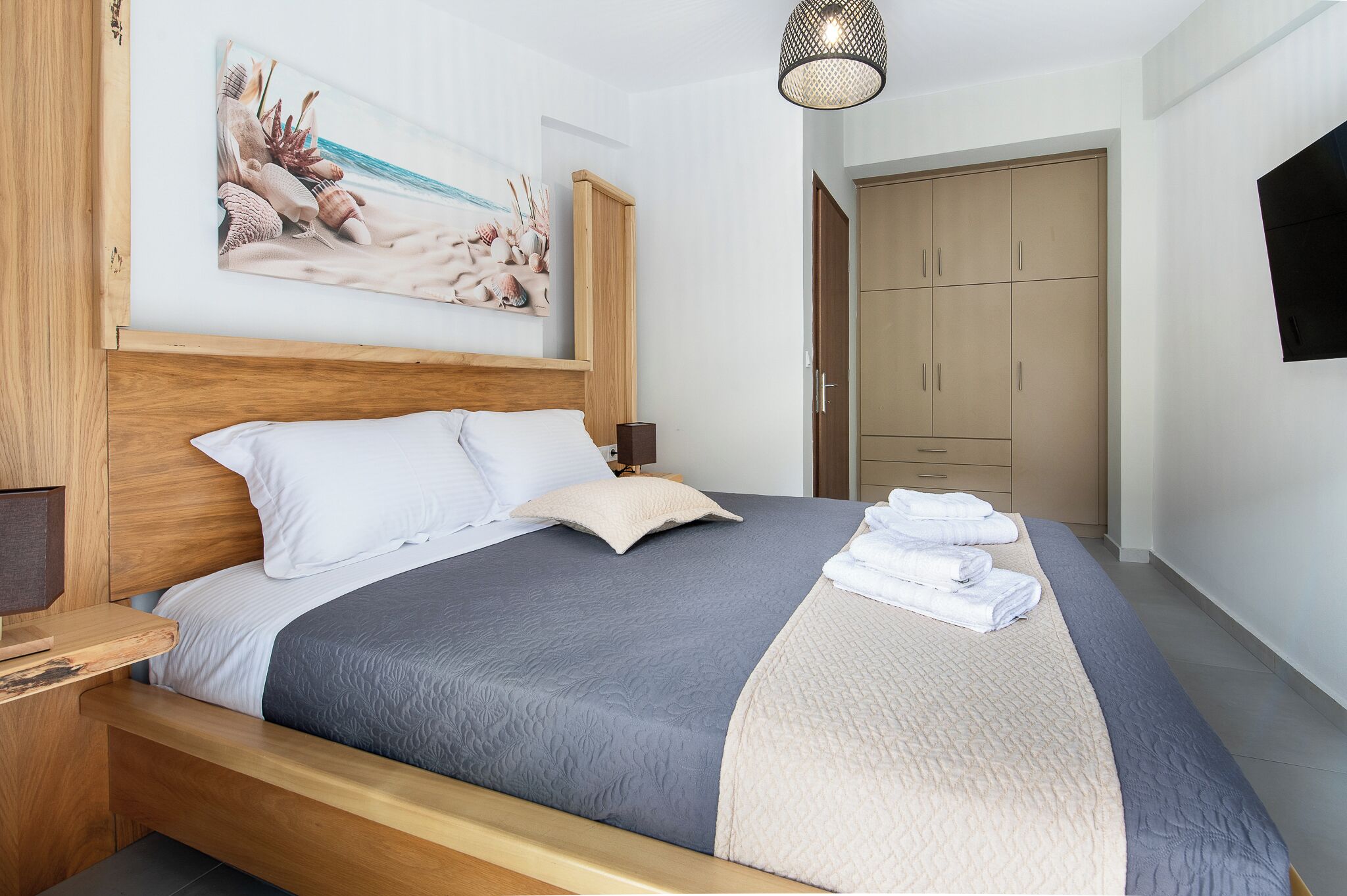 Un lit bien fait, recouvert d’une couverture bleue et beige, avec une tête de lit en bois et un tableau représentant une scène de plage accroché au mur.