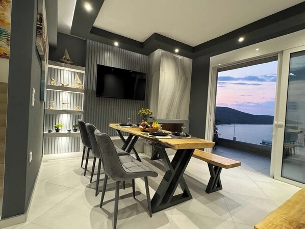 A modern dining area with a long wooden table, four grey chairs, and a bench. A flat-screen TV is mounted on the wall, and there is a view of a body of water through a large sliding glass door. - Suite 3 on the Hill - Stunning, Spacious, and Breathtaking Panoramic View (Amfilochia)