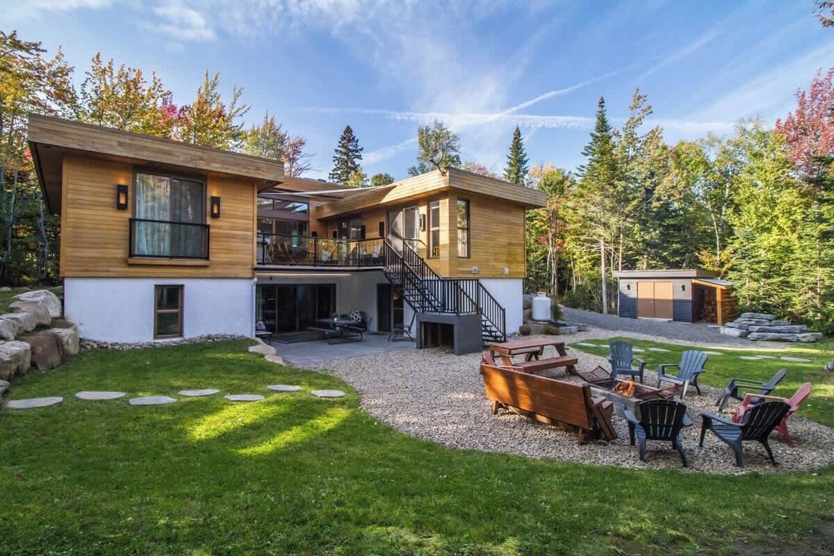 Une maison moderne à deux étages, dotée d’un espace extérieur aménagé avec un coin salon couvert, d’un escalier et d’un petit abri de jardin.