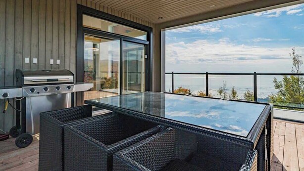 A balcony with a glass-top table and wicker chairs, a grill, and a view of the ocean.