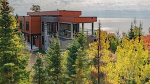 A modern two-story house with a wooden exterior, large windows, and a balcony overlooking a body of water.