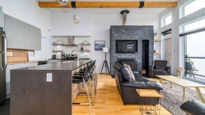 A modern kitchen with a central island, a dining area with chairs, a black leather sofa, and a fireplace.