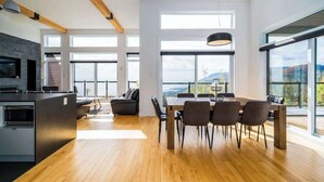 A modern kitchen with a large island, dining area with a wooden table and chairs, and large windows offering views of the outdoors.