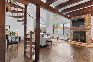 A spacious living room with a wooden staircase, a stone fireplace, and large windows overlooking a body of water.