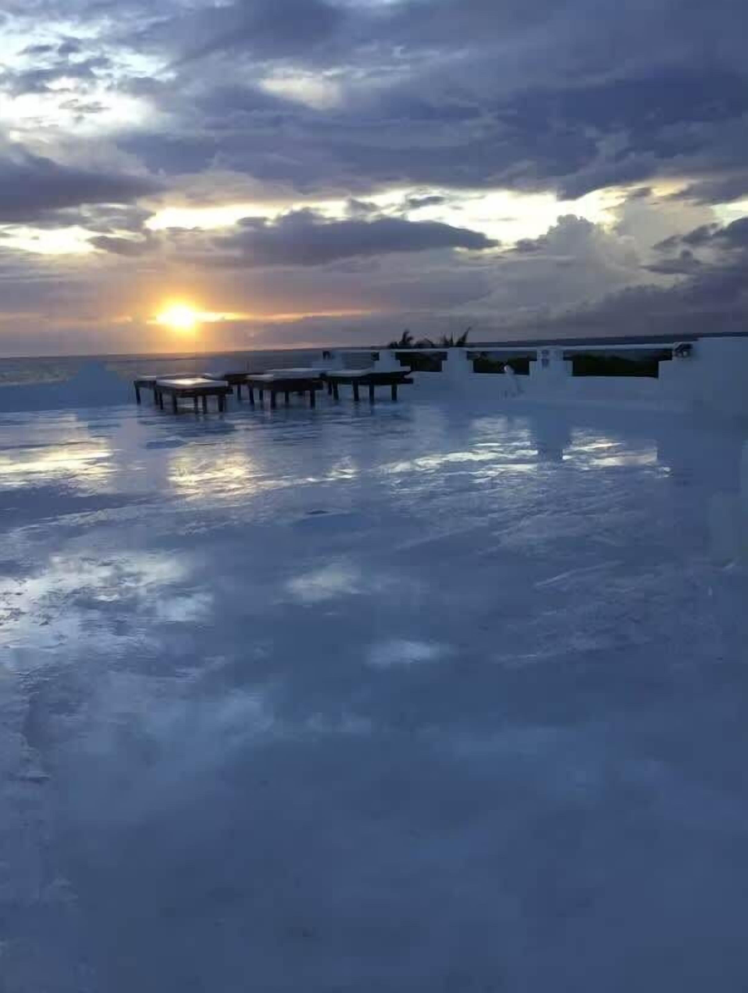 A beachside view with a sunset, a pier, and lounge chairs.