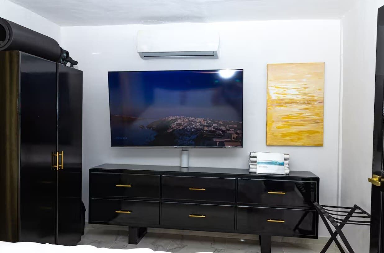 Une chambre moderne avec un grand téléviseur à écran plat fixé au mur, une commode noire avec des poignées dorées et un grand tableau abstrait.