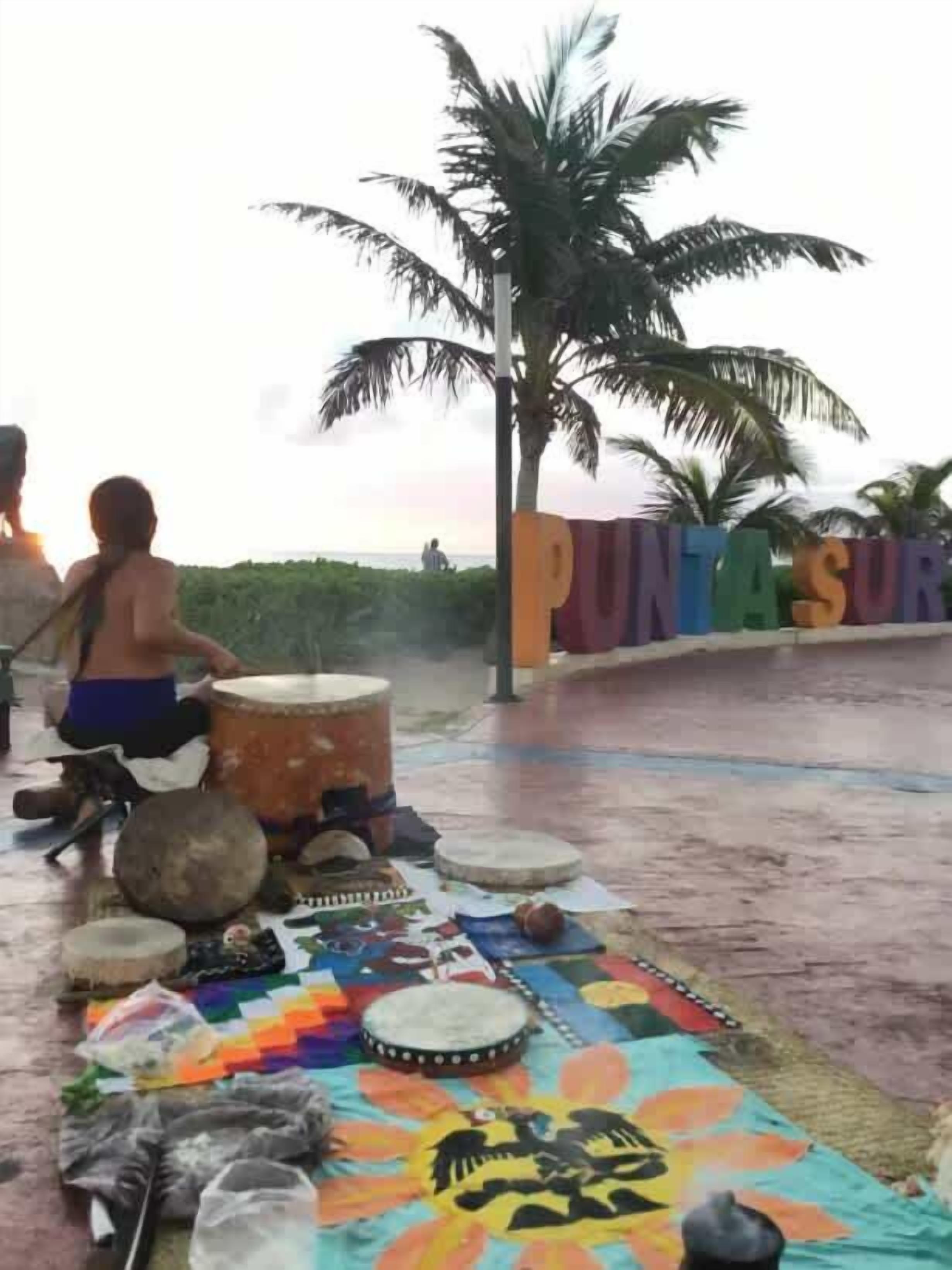 A group of people playing drums outdoors, with a palm tree and a colorful mat on the ground.
