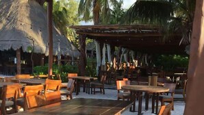 An outdoor seating area with wooden tables and chairs, surrounded by palm trees and a thatched-roof structure. - Luxury One Minute Beach Pool Soak Fast WIFI Villa (Isla Mujeres)