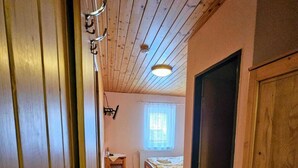 Een smalle gang met houten wanden en een schuine plafond. Aan het einde van de gang staat een bed met een raam en een stoel.