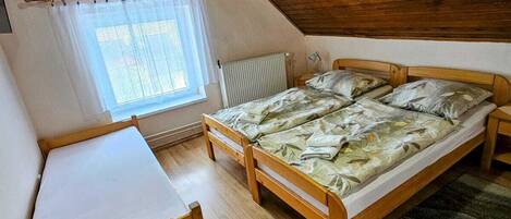 A small, cozy attic bedroom with wooden walls and a sloped ceiling. It features two wooden beds with matching headboards, a small wooden desk with a chair, and a window with a white curtain.
