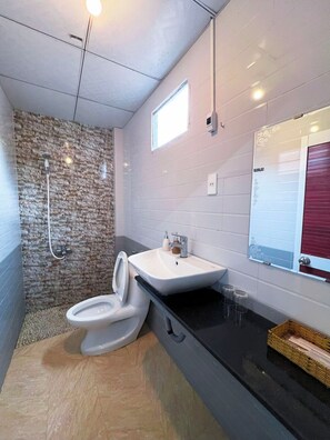 A modern bathroom with a stone shower wall, a white toilet, a sink with a mirror, and a small window. - Sea Breeze Resort & Spa (Phu Quoc)