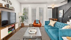 A modern living room with a large flat-screen TV, a blue sofa, a coffee table, and a staircase.