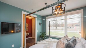 A bedroom with a bed, a mounted TV, a plant, and a window with blinds.