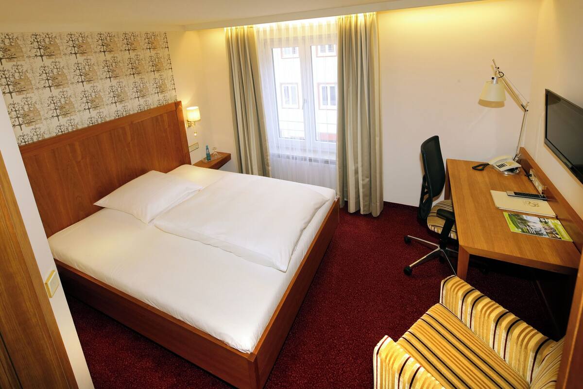 A hotel room with a large bed, a desk with a chair, a TV, and a window with curtains.