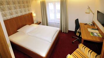 Ein Hotelzimmer mit einem großen Bett, einem Schreibtisch mit Stuhl, einem Fernseher und einem Vorhangfenster.