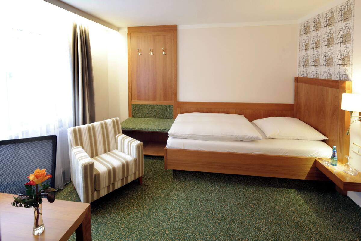 A hotel room with a bed, a chair, a small table with a vase of flowers, a wooden wardrobe, and a wall with a patterned design.