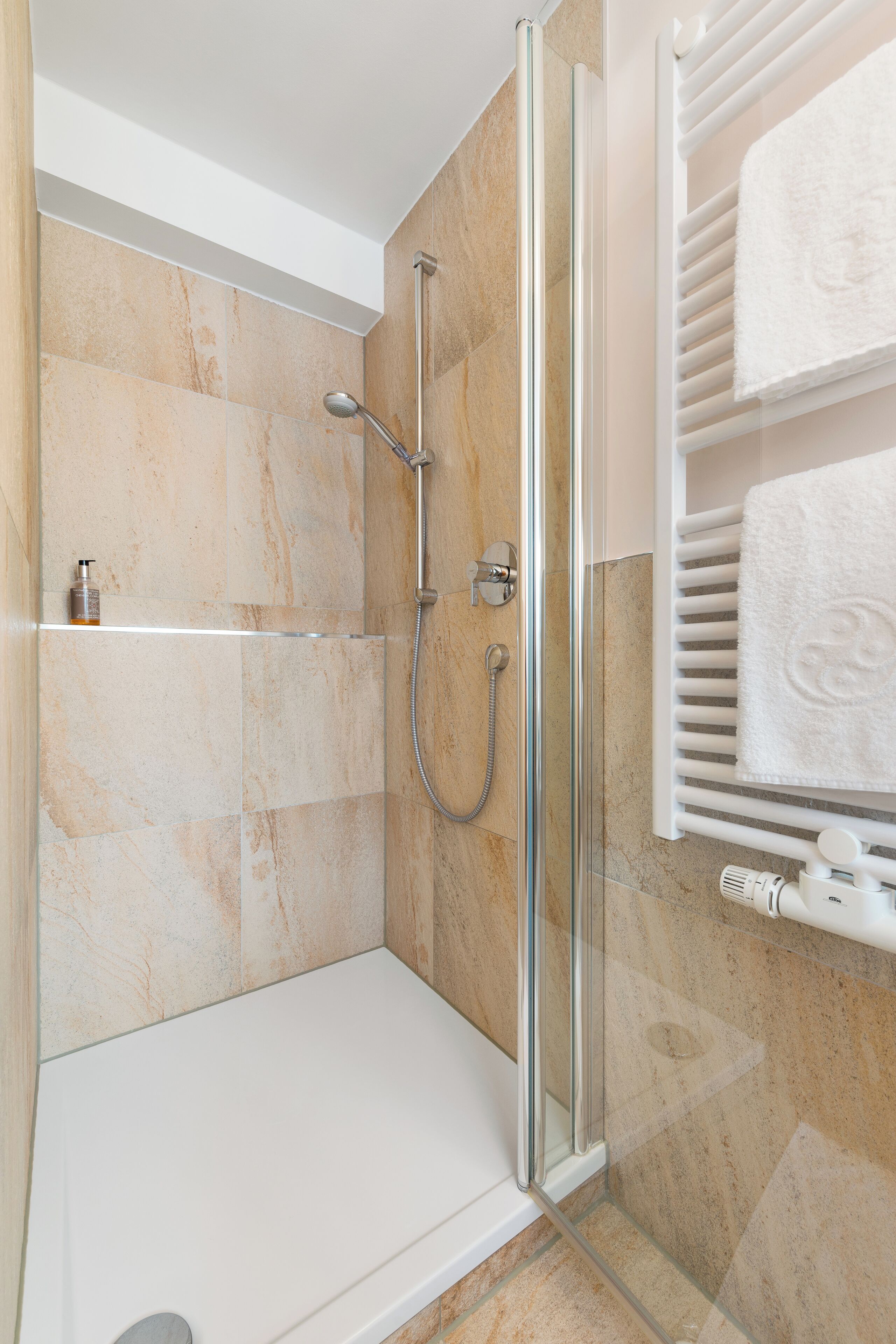 A modern shower with a glass door, beige tiles, and a white towel rack with towels.