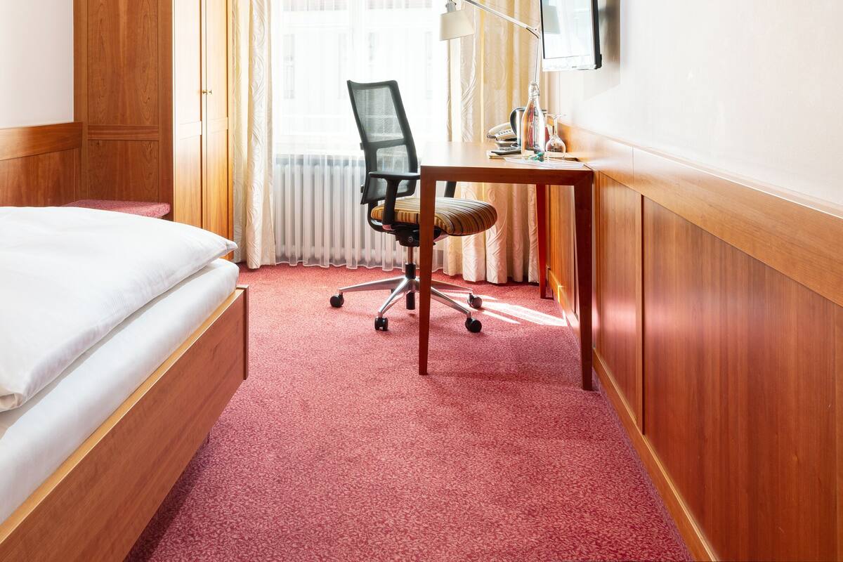 A hotel room with a bed, a desk with a chair, a lamp, and a window with curtains.