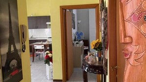 A hallway with a wooden door, a mirror, and a table with flowers. There is a kitchen in the background with cabinets and a fan.