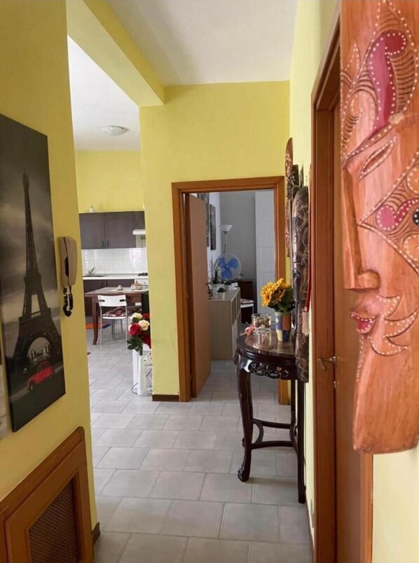 A hallway with a wooden door, a mirror, and a table with flowers. There is a kitchen in the background with cabinets and a fan.