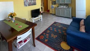 A living room with a sofa, a TV, a dining table with a vase of flowers, and a kitchen area with cabinets and a refrigerator.