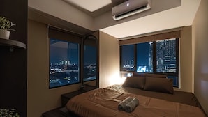 Una habitación de hotel moderna con una cama grande, dos ventanas con vistas a la ciudad, una mesita de noche pequeña y un equipo de aire acondicionado.