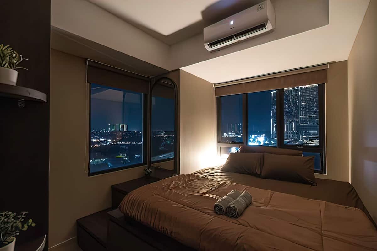 Una habitación de hotel moderna con una cama grande, dos ventanas con vistas a la ciudad, una mesita de noche pequeña y un equipo de aire acondicionado.
