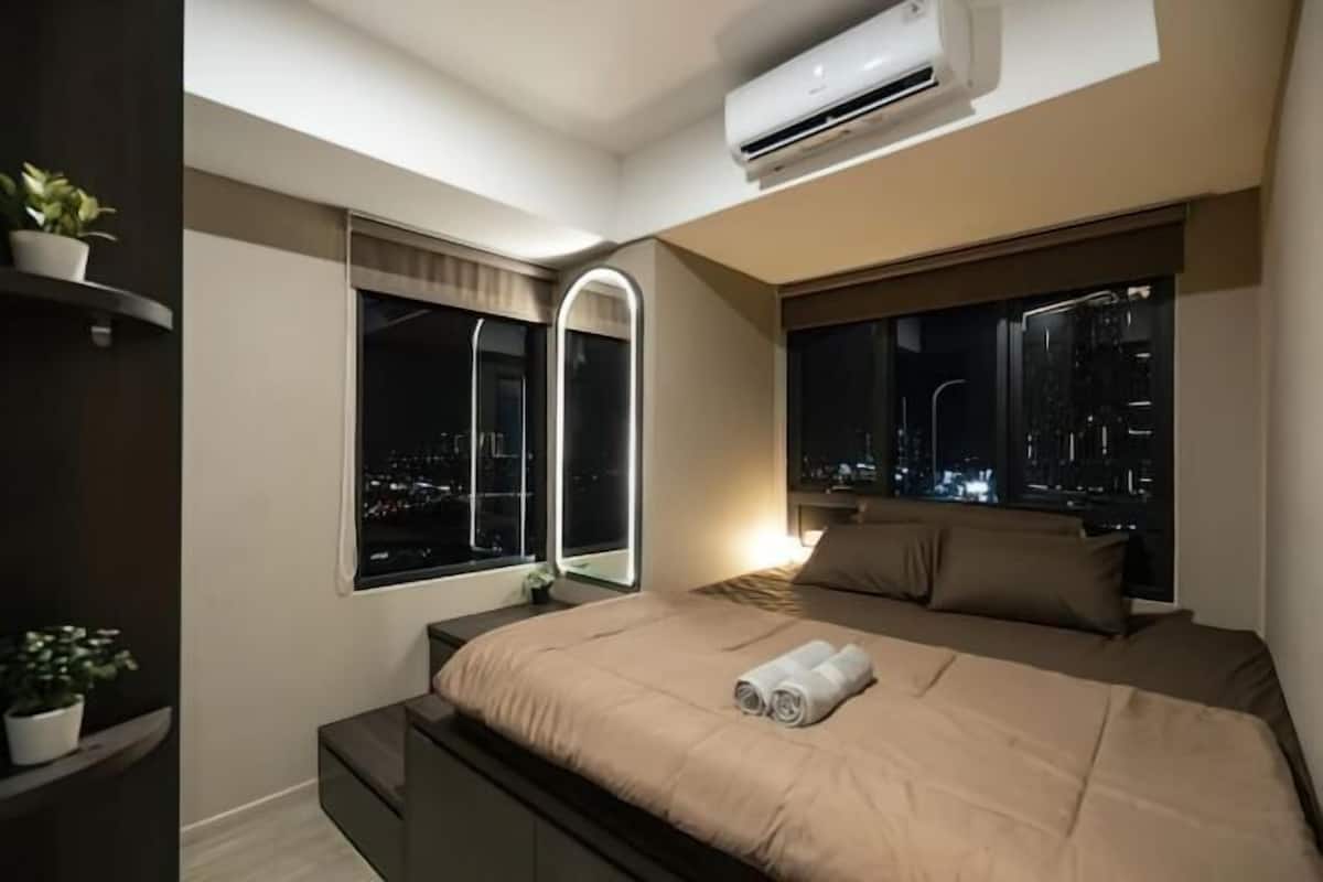Un dormitorio moderno con cama, mesitas de noche, unidad de aire acondicionado, ventana con vista a la ciudad de noche y un espejo.