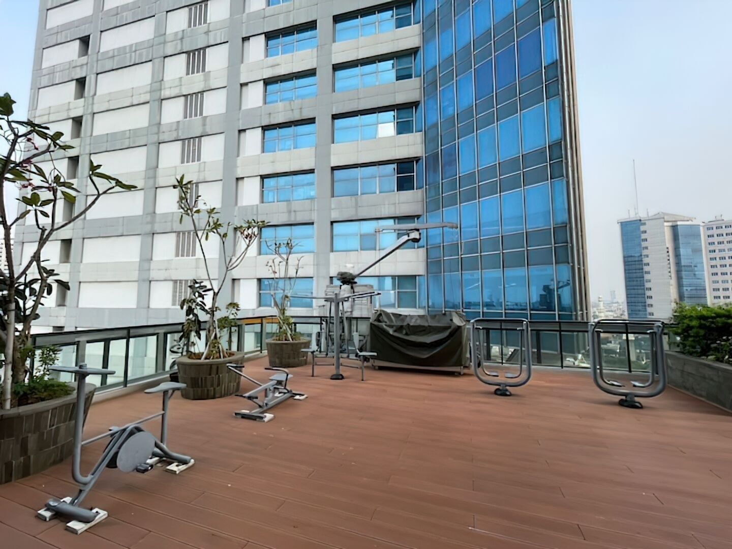 Terraza en la azotea con equipo de ejercicio, plantas en macetas y vistas a un edificio moderno.