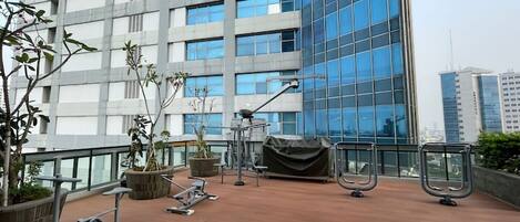 Terraza en la azotea con equipo de ejercicio, plantas en macetas y vistas a un edificio moderno.