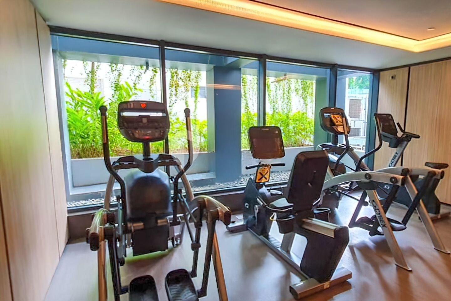 A modern gym with various exercise equipment, large windows with a view of greenery, and a clean, well-lit interior.