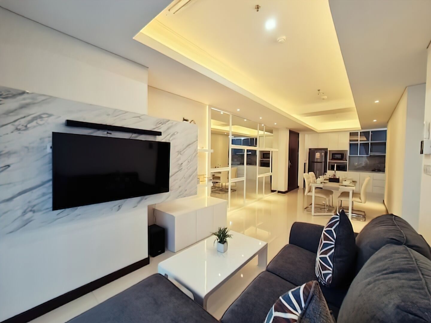 A modern living room with a flat-screen TV mounted on a marble wall, a sofa, a coffee table, and a dining area in the background.