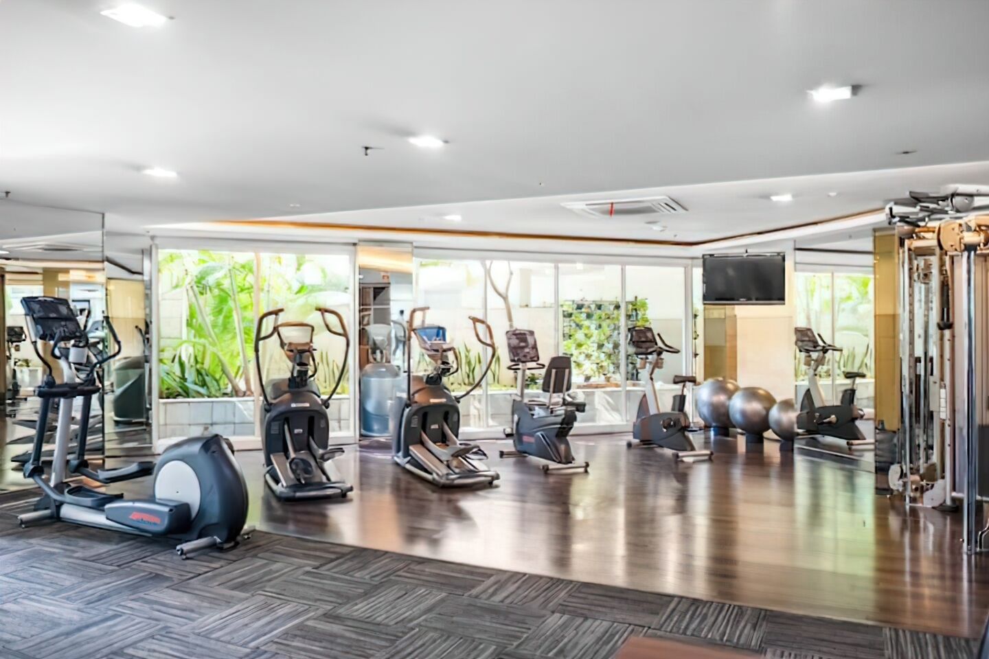 Une salle de sport moderne équipée de divers appareils de cardio, de miroirs et d’une grande fenêtre donnant sur un espace verdoyant.
