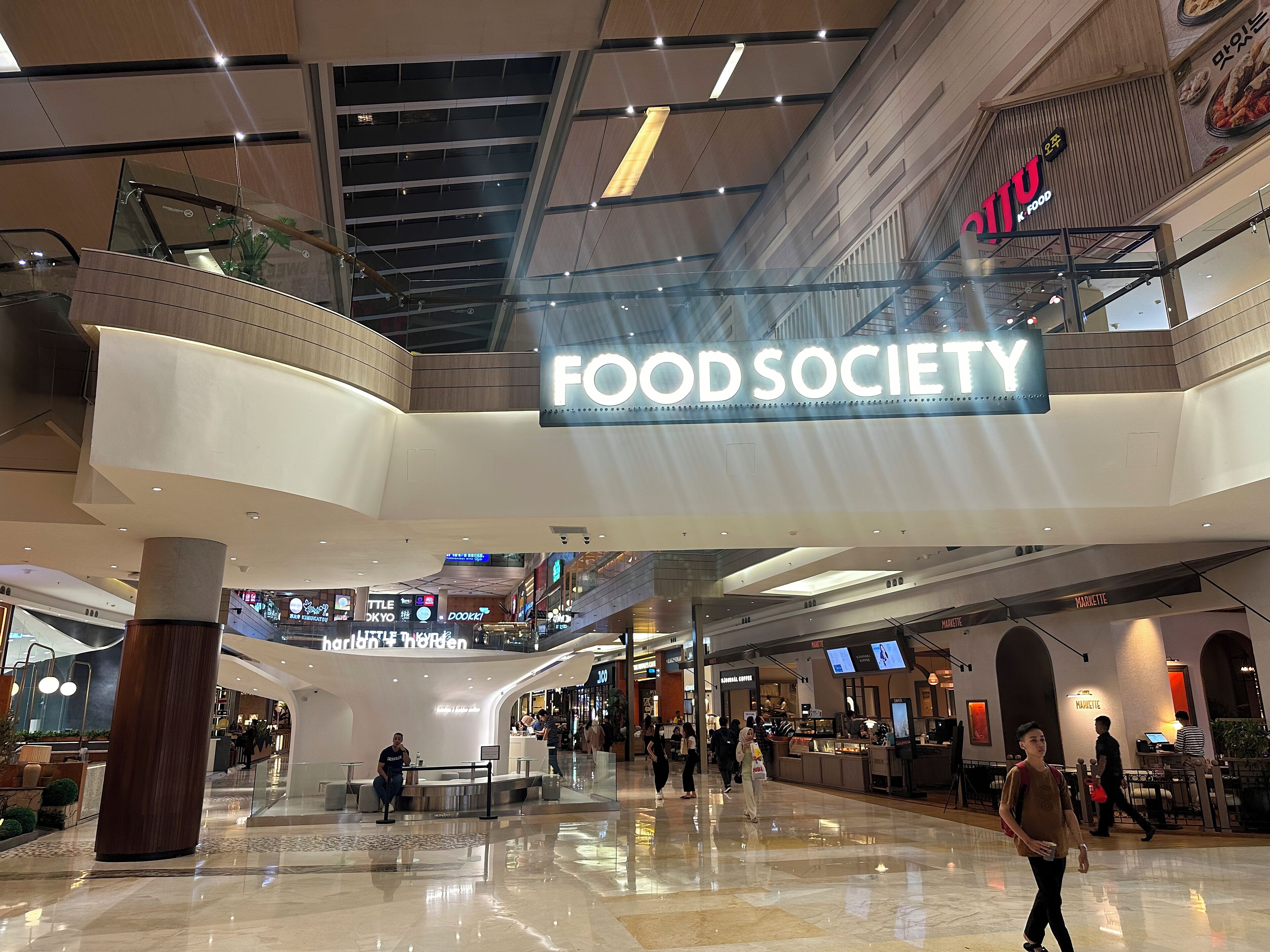 A spacious food court area with a prominent 'FOOD SOCIETY' sign, various food outlets, and a modern architectural design with glass railings and polished floors.