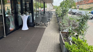 An outdoor seating area with tables and chairs, a potted plant, and a pathway leading to a building entrance. - HOTEL BONÉ TO (Salzgitter)