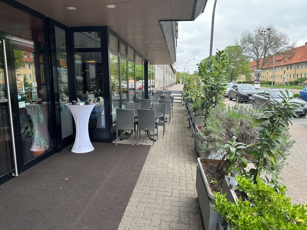 An outdoor seating area with tables and chairs, a potted plant, and a pathway leading to a building entrance. - HOTEL BONÉ TO (Salzgitter)
