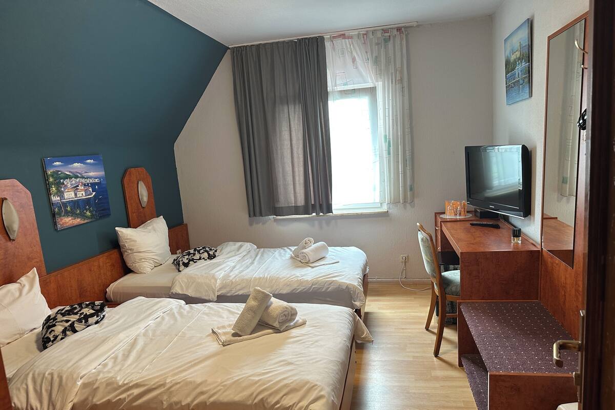 A hotel room with two beds, a desk with a television, a window with curtains, and a painting on the wall.