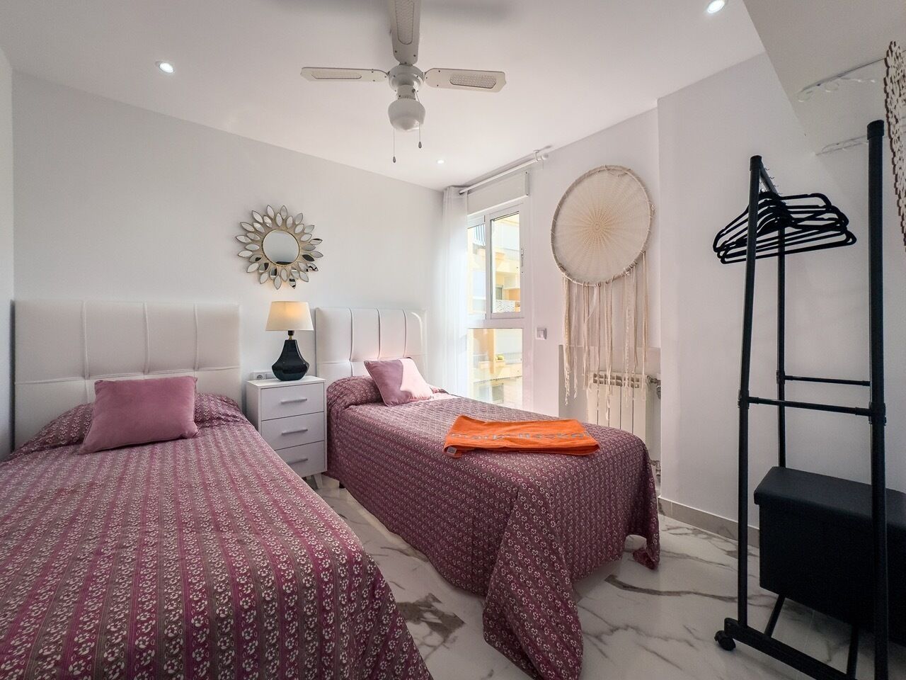 A bedroom with two beds, a ceiling fan, a nightstand, a lamp, a dreamcatcher, and a black shelf.