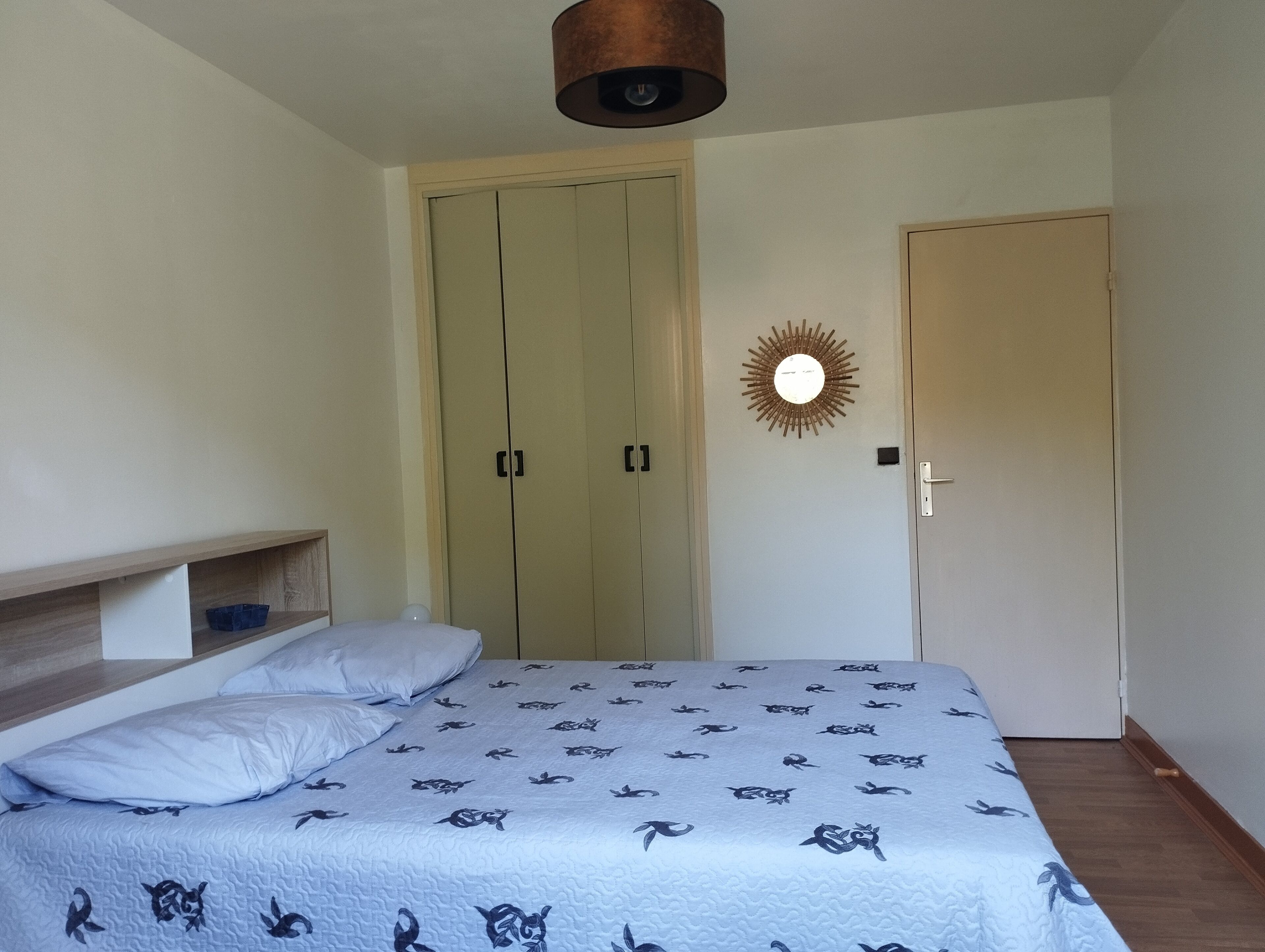 A bedroom with a bed, a wardrobe, a shelf, and a wall-mounted light fixture.