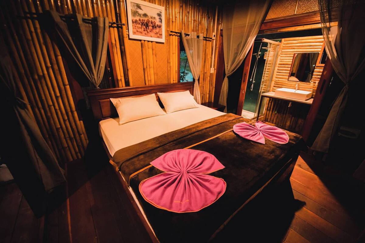 A wooden-framed room with a bed, a bathroom with a mirror, and bamboo walls.