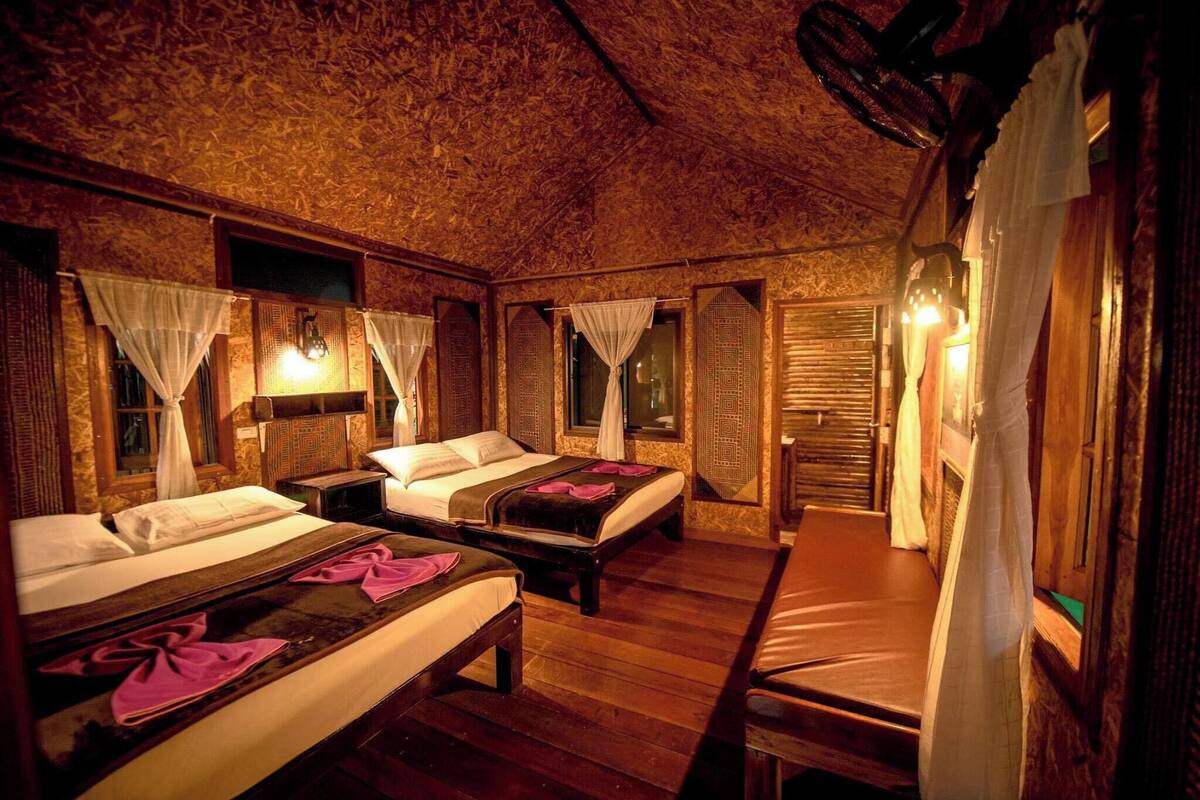 A room with three wooden beds, each with white linens and pink towels. There are wall-mounted lights and a wooden ceiling.