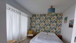 Un dormitorio con cama, una silla, una mesita, un radiador y una pared con estampado de hojas tropicales.