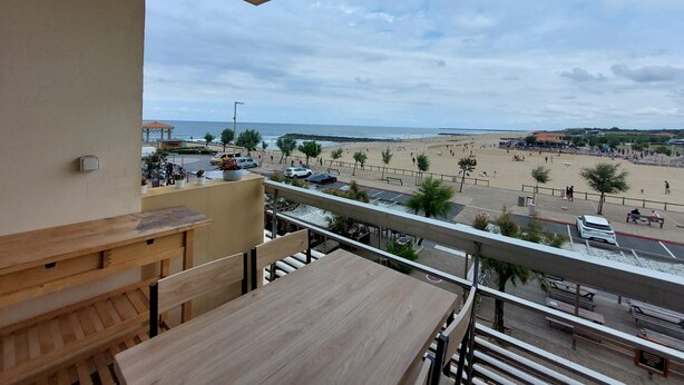 Un balcón con mesa y sillas de madera, con vista a una playa donde hay gente y vehículos.