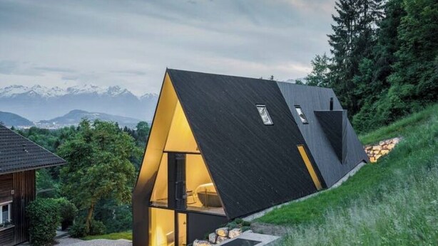 Une maison moderne, de forme triangulaire, avec un toit en pente, de grandes fenêtres et une vue sur les montagnes en arrière-plan.