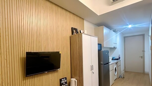 Une chambre d’hôtel moderne avec un mur revêtu de panneaux de bois, un téléviseur à écran plat fixé au-dessus d’une console en bois, un lit avec une couette à motifs gris, une kitchenette équipée d’un réfrigérateur et d’un lave-linge, ainsi qu’un petit coin repas.